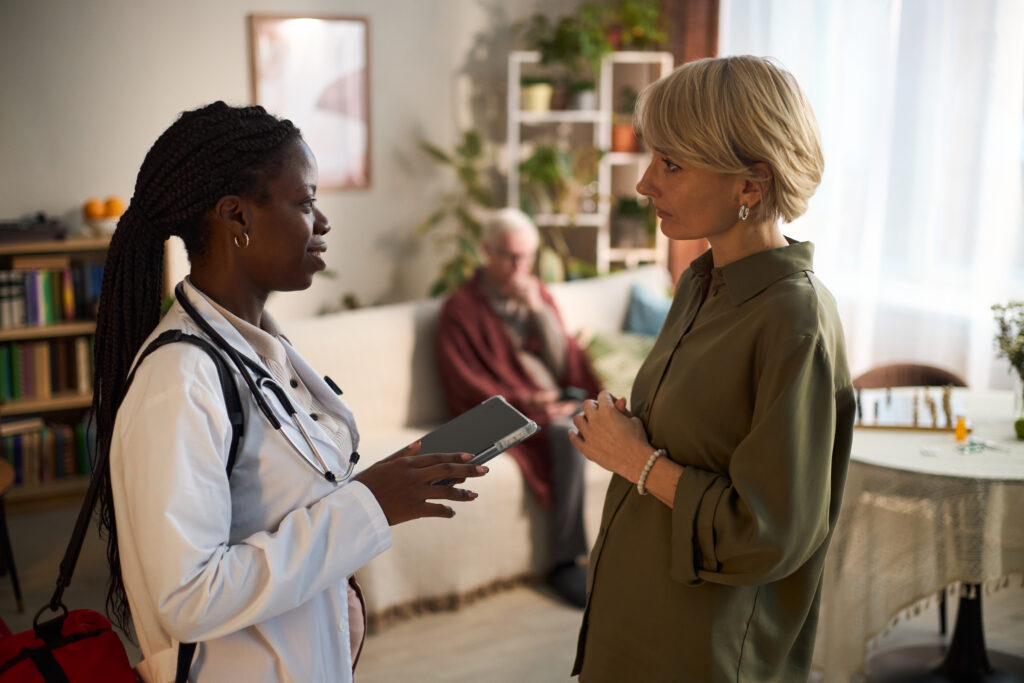 Black young adult woman doctor holding digital tablet, speaking with Caucasian middle aged woman in living room, while senior Caucasian man sitting on sofa in background